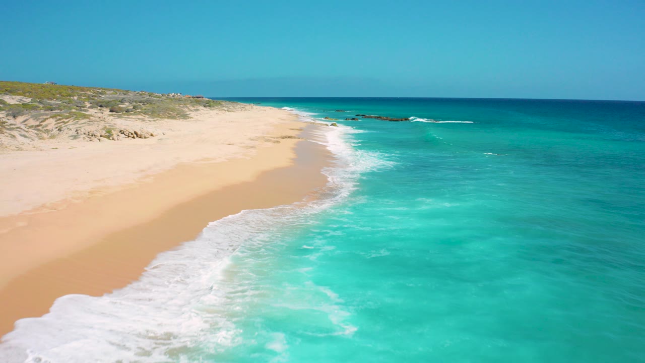 Aerial frame from El Cardoncito, Baja California Sur showing turquoise water washing onto a wide sandy beach with low dunes behind it and no visible people.