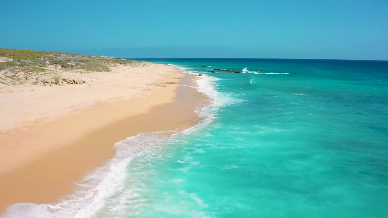 Aerial frame from Baja California Sur showing bright turquoise shallows, a narrow white wave break, dry sand to the left, and no visible people.