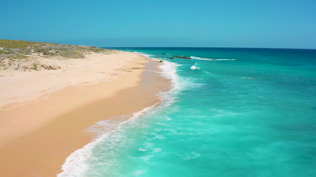 Elevated El Cardoncito shoreline view with white surf entering from the lower left, open sand through the center, blue water to the right, and no visible people.