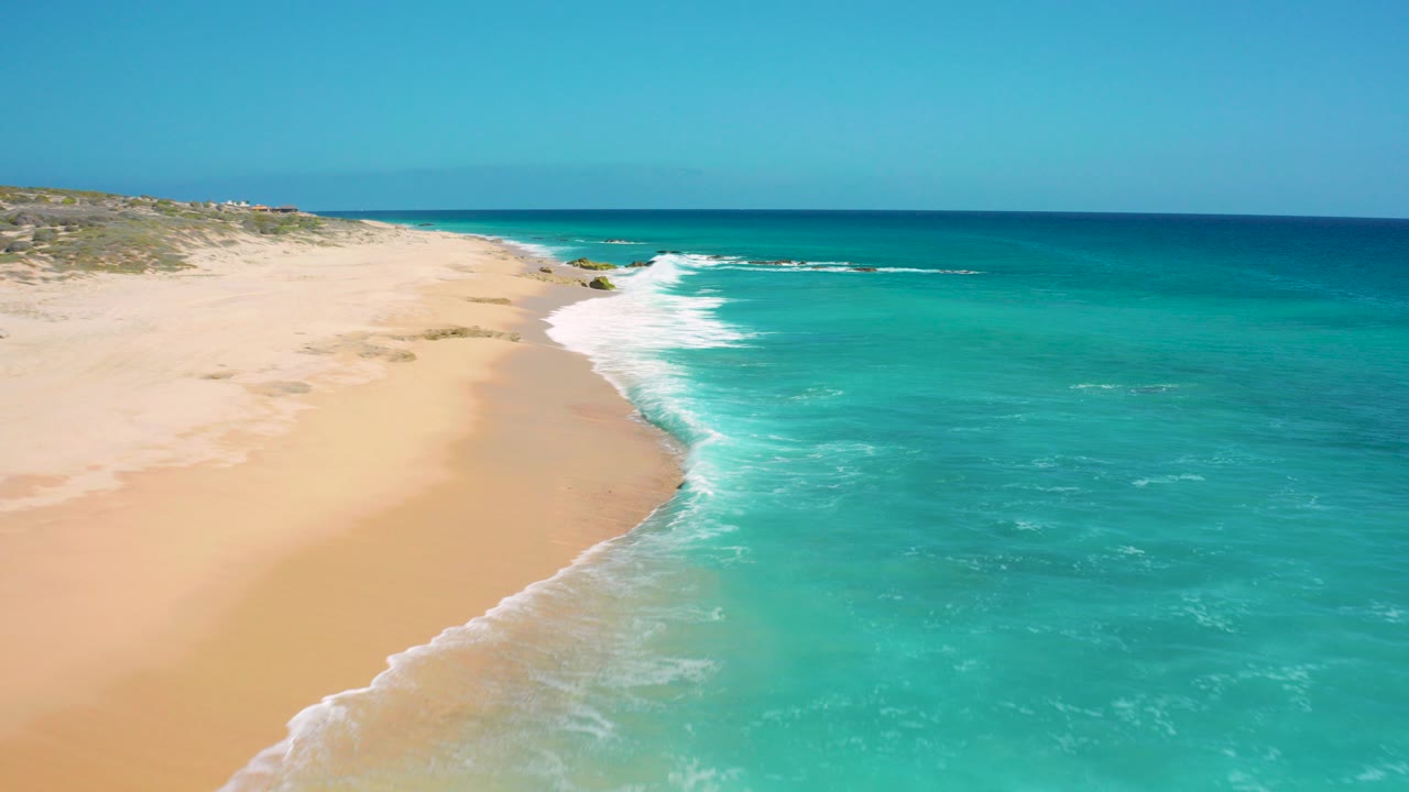 Aerial view at El Cardoncito, Baja California Sur, with pale sand on the left, turquoise water on the right, white surf curving along the shore, low dune vegetation in the distance, and no visible people.
