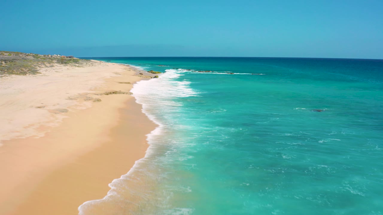 At 0:02, the aerial composition shifts slightly along the El Cardoncito coast, showing a stronger band of white surf against the sand, deeper blue water beyond, and no visible people.
