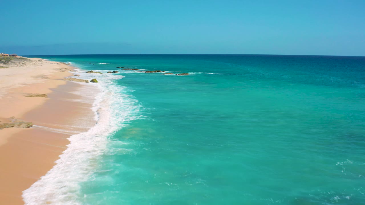 El Cardoncito beach seen from above with tan sand narrowing at the left edge, a broad turquoise sea filling the frame, small rocks near the upper surf, and no visible people.