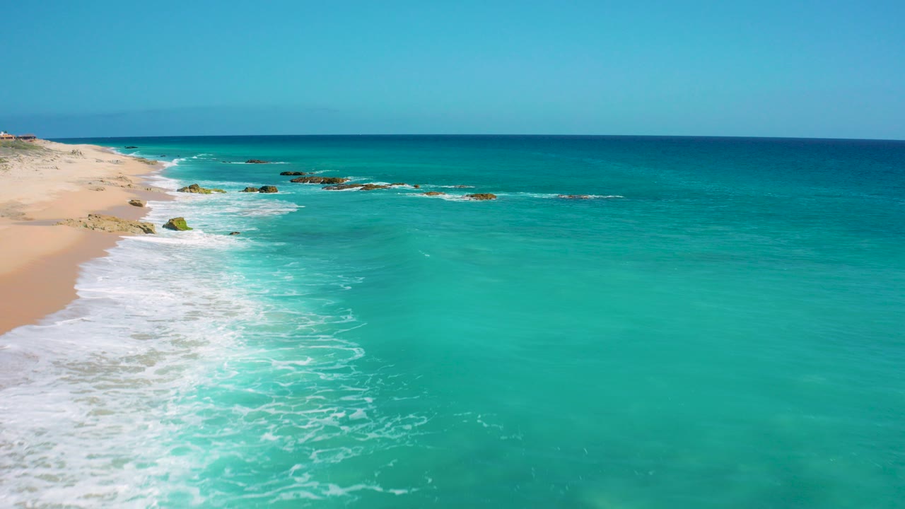 Opening aerial frame over El Cardoncito shows white surf curling along the sandy beach at left, turquoise water across the foreground, distant rocks near the break, and no visible people.