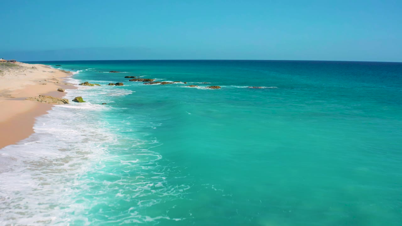 At 0:01, the aerial view remains over El Cardoncito with the surf line slightly farther down the left edge, clear blue-green water dominating the frame, small rocks offshore, and no visible people.