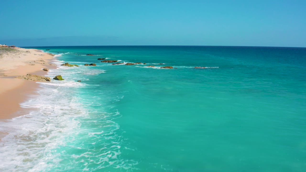 At 0:02 in El Cardoncito, turquoise ocean fills the right side while white foam spreads along the sandy shore at left, with scattered rock patches near the wave break and no visible people.