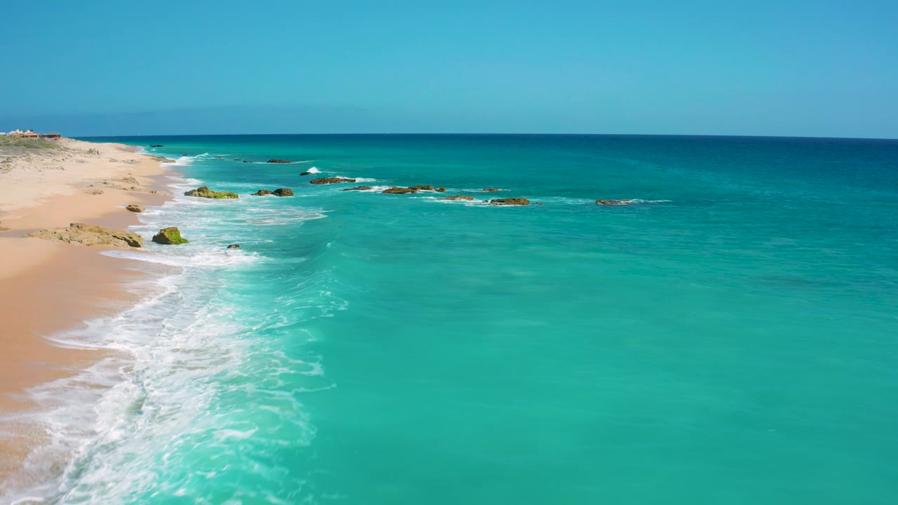 At 0:03, the El Cardoncito beach curves through the left side with white waves breaking against pale sand, open turquoise water in the foreground, low rocks near shore, and no visible people.