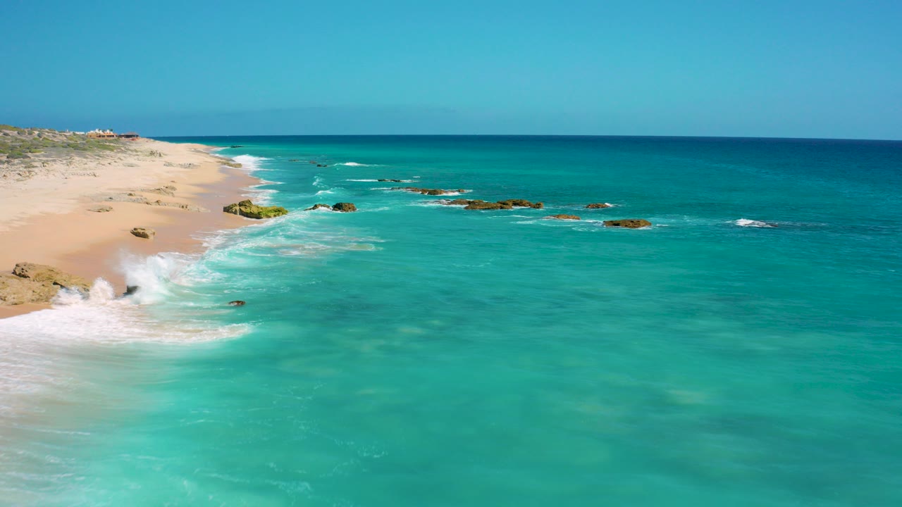 Aerial view over El Cardoncito, Baja California Sur, with turquoise water, pale sand along the left edge, scattered rocks in the shallows, a clear horizon, and no visible people.