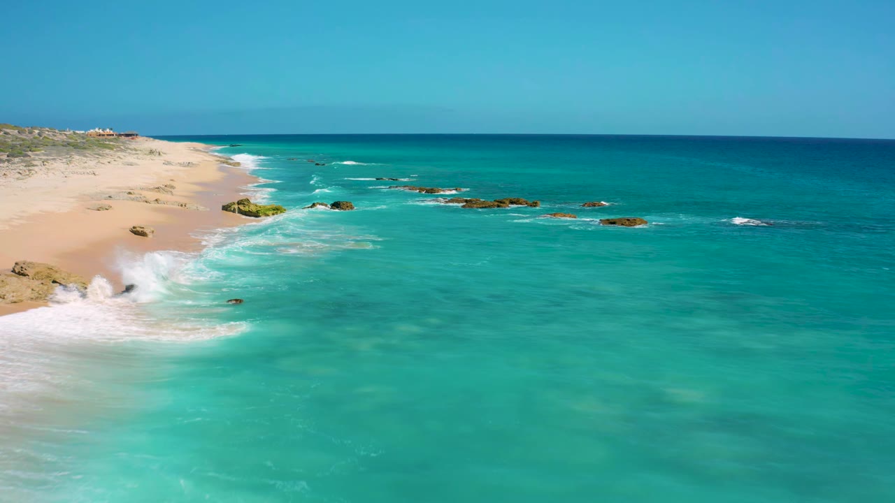 Wide aerial frame of El Cardoncito coastline where white waves curl onto the pale beach, dark rocks sit offshore, the sea shifts from aqua to deep blue, and no visible people appear.