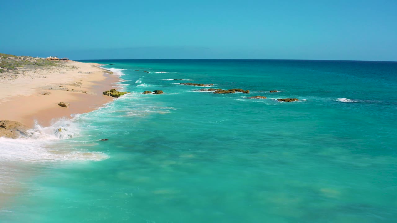Aerial view of El Cardoncito with the sandy beach narrowing into the distance, turquoise water across the foreground, rocks near the surf zone, a flat blue horizon, and no visible people.