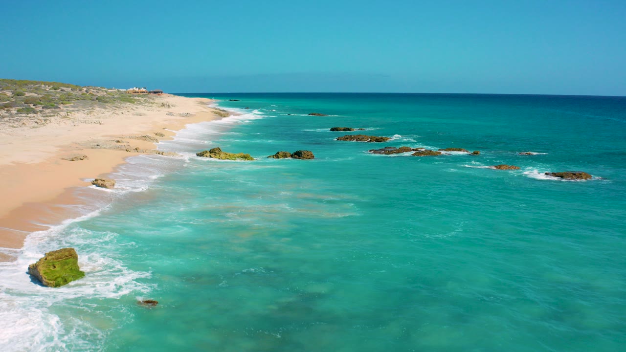 Aerial frame over El Cardoncito, Baja California Sur showing turquoise water washing toward a pale sandy beach with scattered rocks and no visible people.