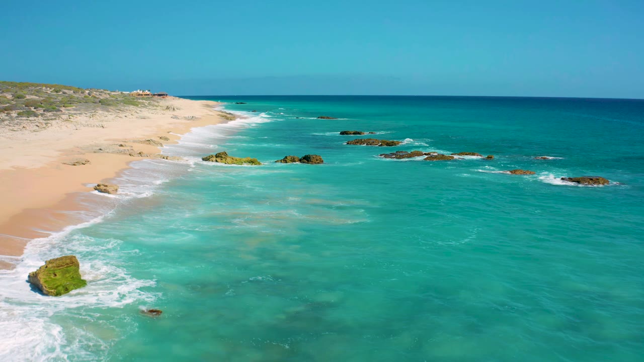 El Cardoncito aerial view with the beach running along the left side, shallow turquoise water across the foreground, dark rocks offshore, and no visible people.