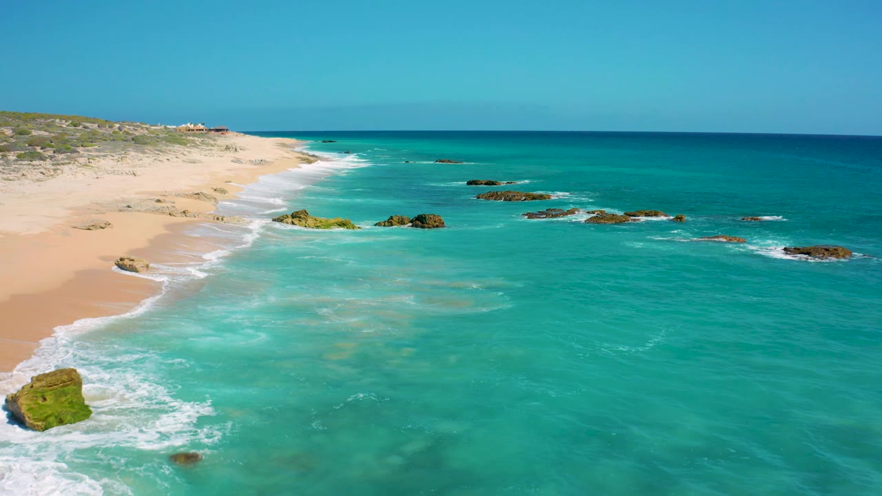Aerial view in Baja California Sur with blue-green water filling most of the frame, a sandy beach at left, rocks in the surf, and no visible people.