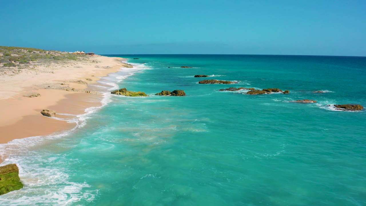 Mid-clip aerial frame over El Cardoncito showing white surf curling along the sand, rocky outcrops in the water, open turquoise sea, and no visible people.