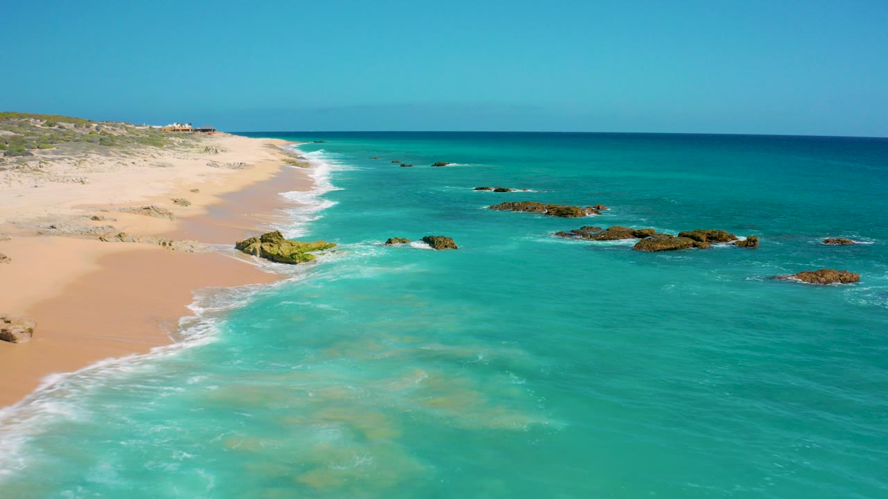 Aerial frame over El Cardoncito, Baja California Sur, showing pale sand on the left, turquoise water on the right, dark rock outcrops offshore, white surf, and no visible people.