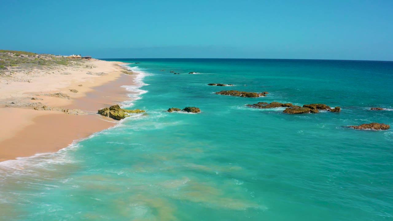 Aerial view at El Cardoncito with more open water across the right side, pale beach and low dunes at left, small surf breaks, dark rocks offshore, and no visible people.