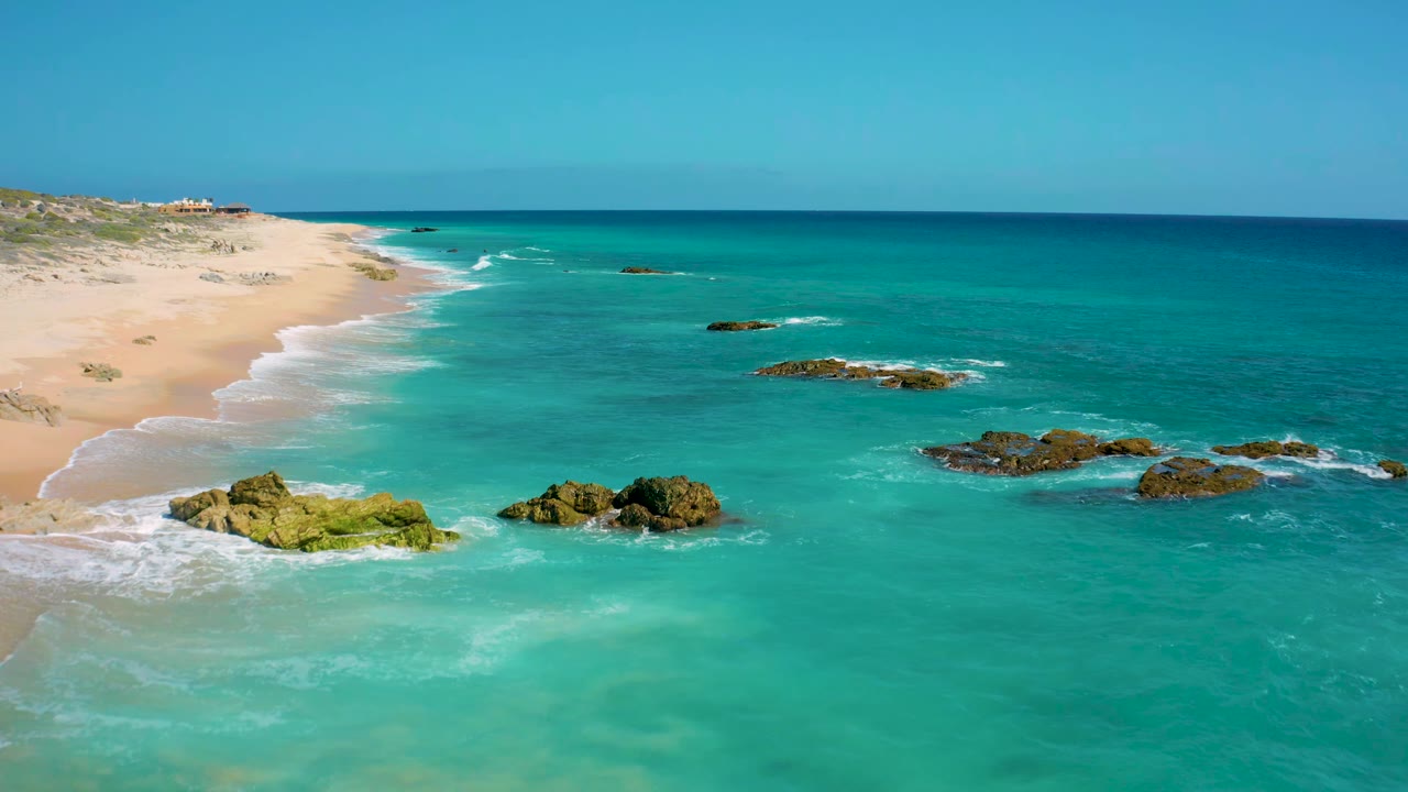 El Cardoncito beach remains on the left as turquoise water fills the frame, with dark reef rocks in the midground, small white wave breaks, distant low hills, and no visible people.