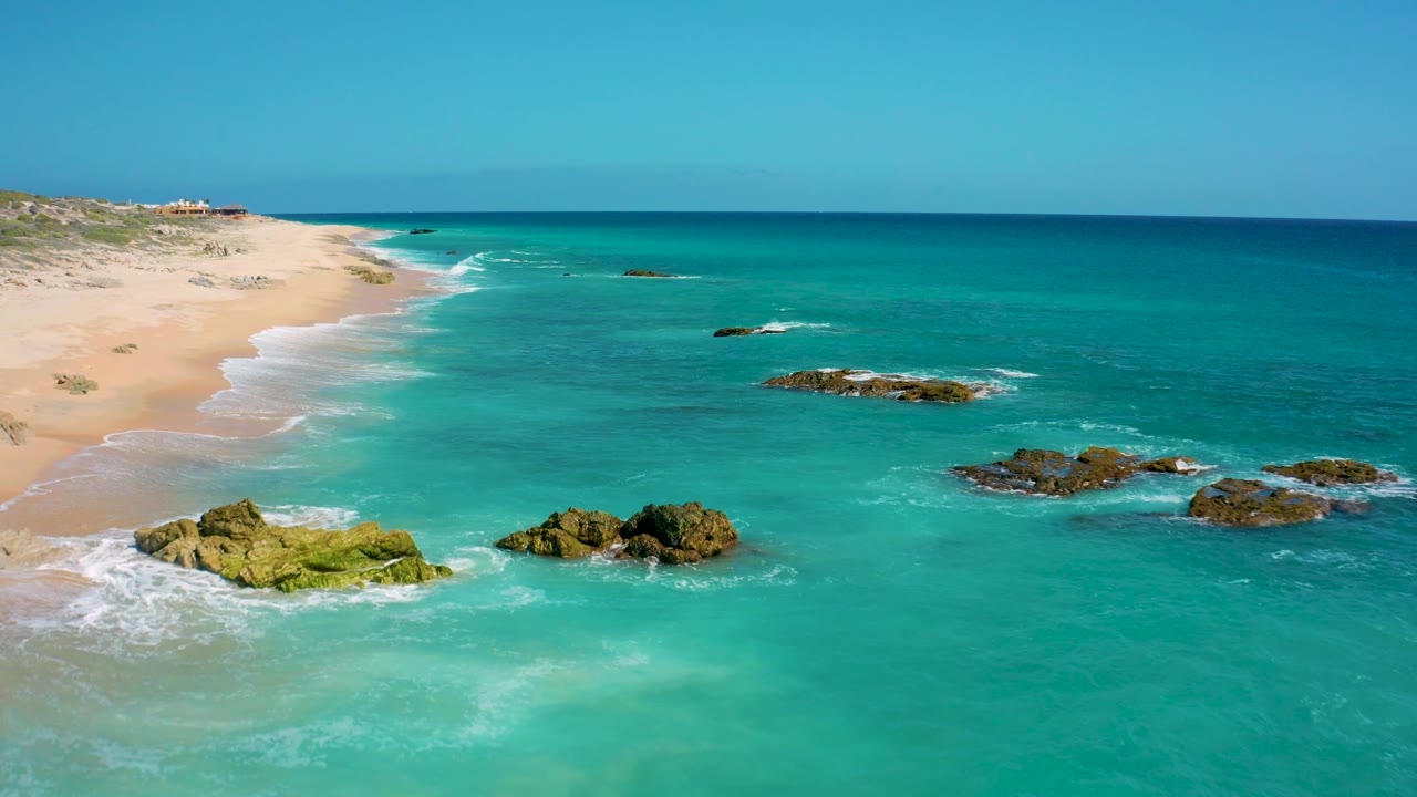 A bright view over El Cardoncito shows blue-green water, reef rocks surrounded by foam, an empty sand beach along the left edge, low vegetation beyond the sand, and no visible people.
