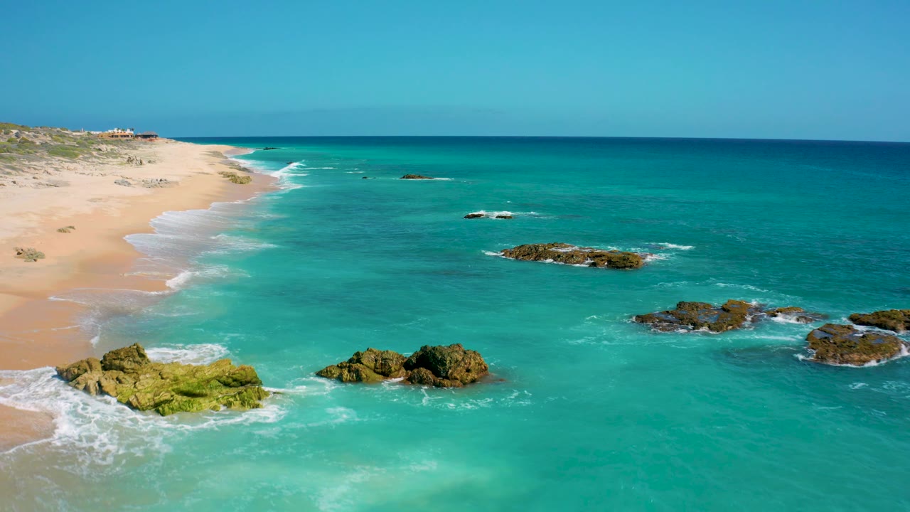 El Cardoncito coastline appears from above with rock clusters near the lower left and right, turquoise water across the center, a long empty beach on the left, and no visible people.