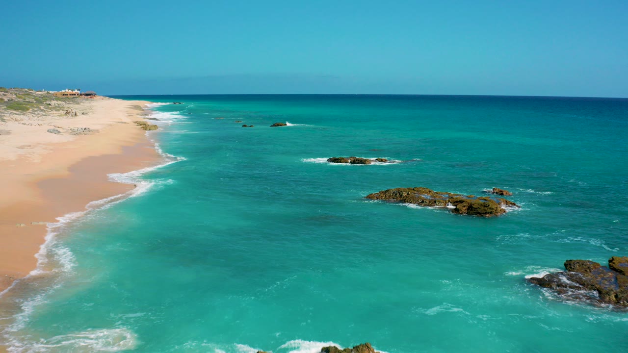 Aerial frame from El Cardoncito, Baja California Sur showing pale sand at left, turquoise water across the center, dark rocks near the lower right, and no visible people.