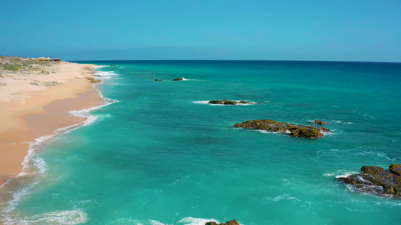 Aerial view over El Cardoncito with the beach running along the left side, small white wave lines on the shore, rocks in the middle distance, and no visible people.