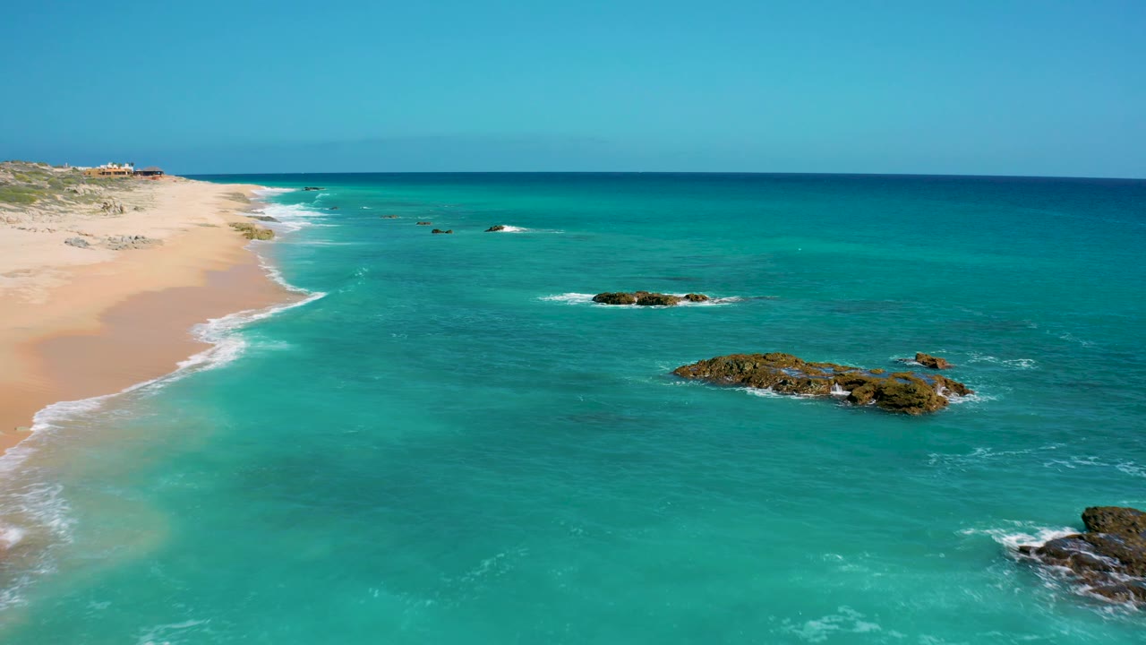 El Cardoncito aerial frame with open blue-green ocean dominating the view, a sandy beach strip at left, exposed dark rocks near center right, and no visible people.