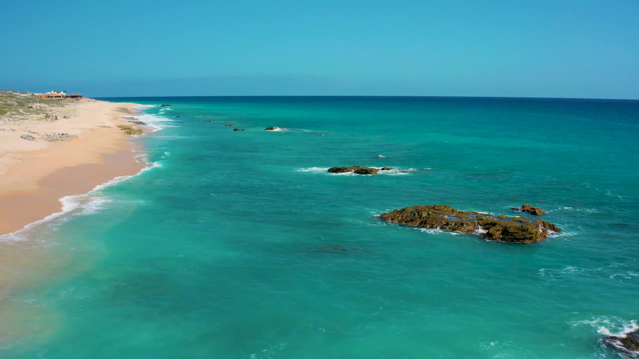 High oblique aerial of El Cardoncito showing the sandy coastline on the left, turquoise water filling most of the frame, a larger rocky outcrop near the lower right, and no visible people.