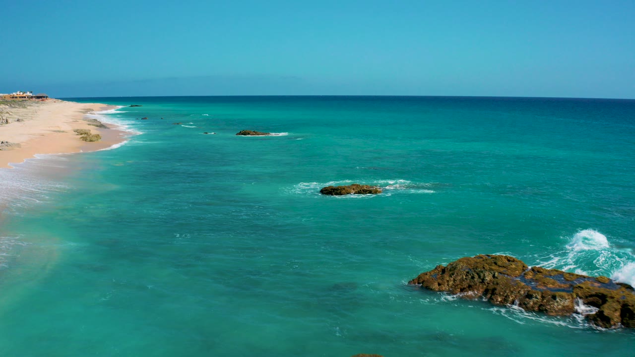 Aerial view at El Cardoncito, Baja California Sur, with turquoise water, pale sand on the left, dark surf rocks near the lower right, a level horizon, and no visible people.