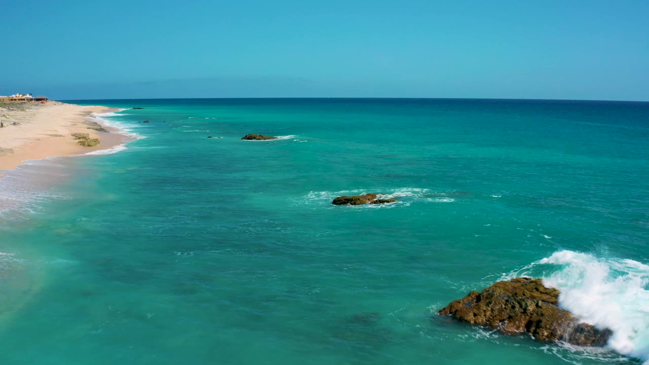 At El Cardoncito, a white wave breaks against a dark rock near the lower right while open turquoise sea and a sandy shoreline fill the rest of the aerial frame, with no visible people.