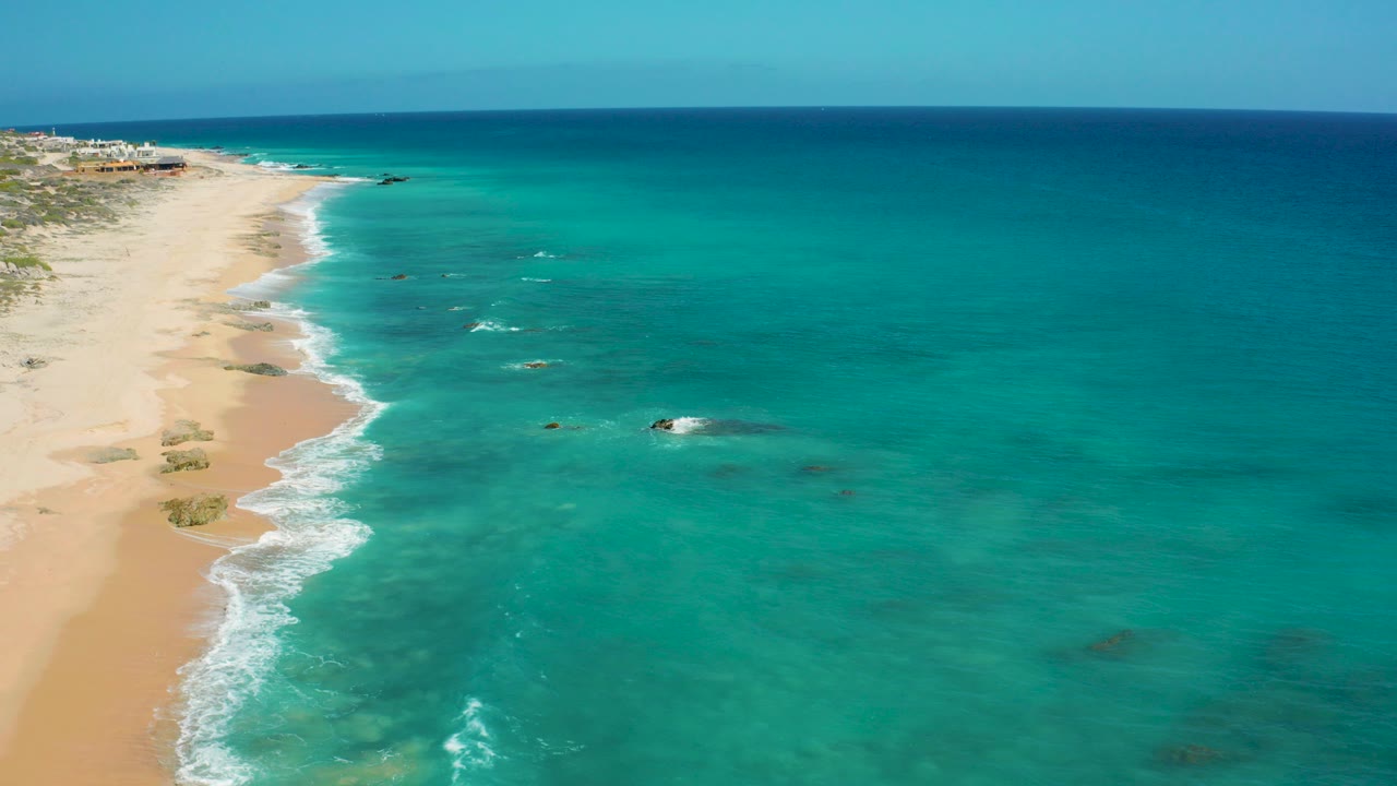 Aerial view at El Cardoncito, Baja California Sur, showing turquoise water beside a sandy beach with low vegetation inland and no visible people.