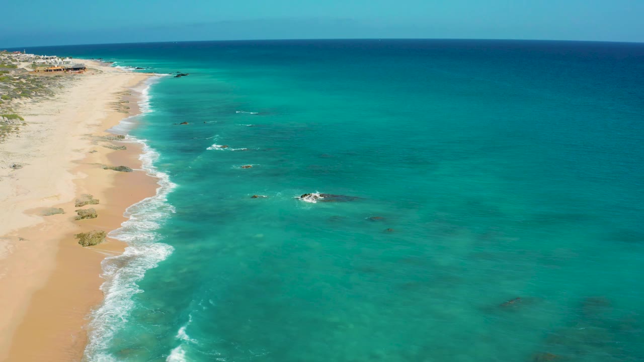 Aerial view at El Cardoncito, Baja California Sur, showing clear water spread across the frame, small reef patches offshore, and no visible people.