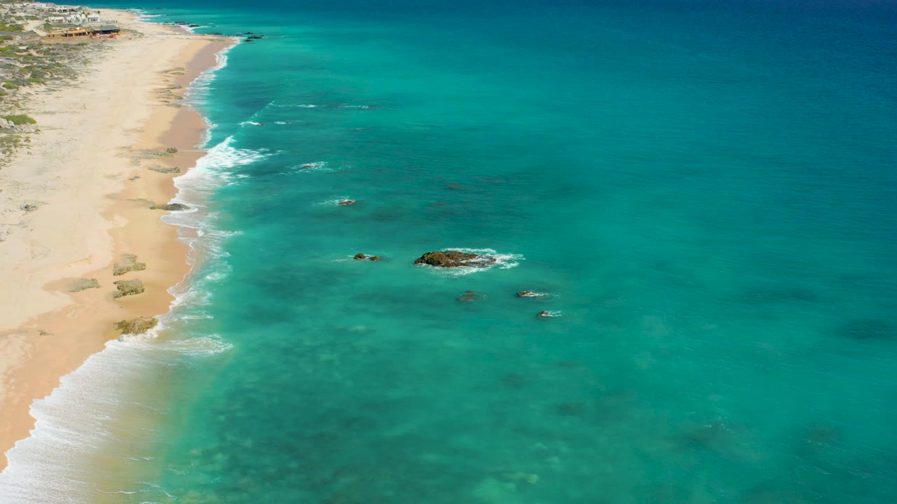 Opening aerial frame over El Cardoncito, Baja California Sur, with pale sand along the left edge, clear turquoise water to the right, small offshore rocks, white surf, and no visible people.