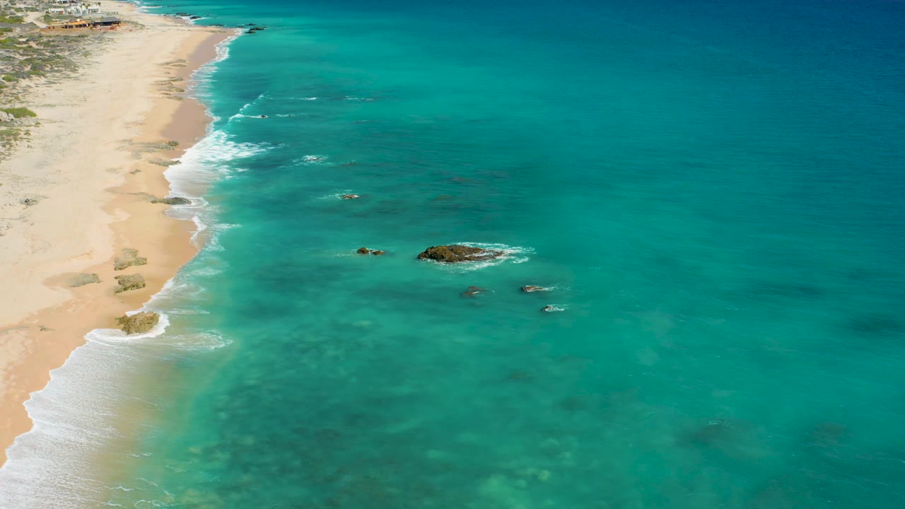 Aerial view over El Cardoncito with the shoreline tight to the left, broad turquoise water filling most of the frame, several dark rocks in the shallows, a calm horizon, and no visible people.