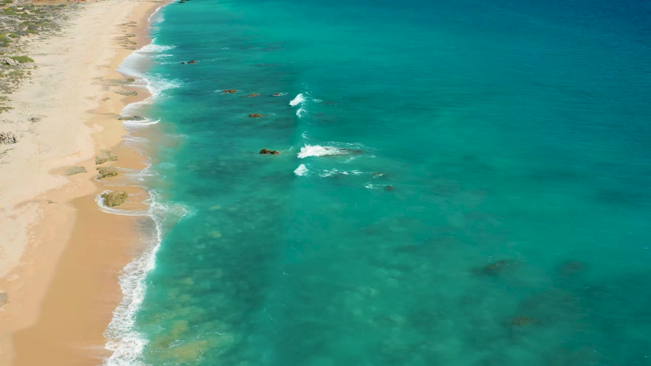 El Cardoncito beach appears from above with the shoreline running diagonally, faint reef color under the water, scattered rocks offshore, and no visible people.