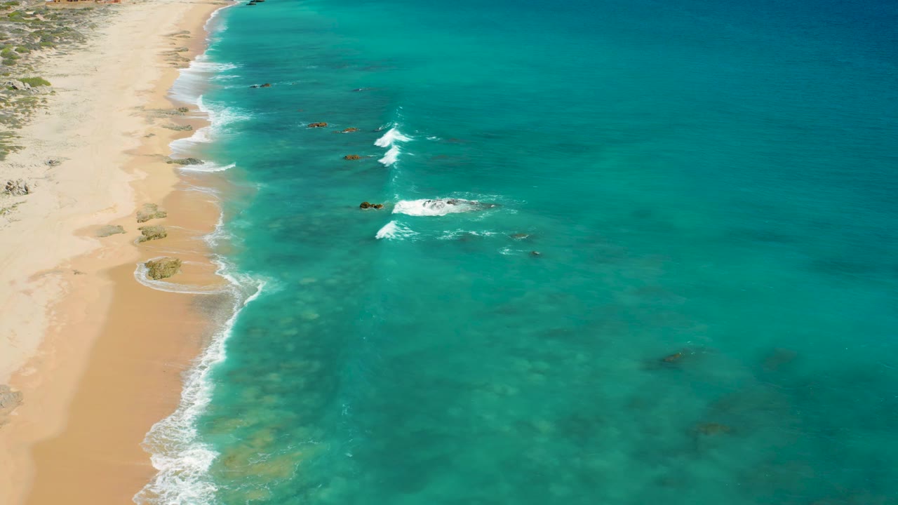 Aerial frame over the Baja California Sur coast with empty sand at left, bright blue-green water at right, foam gathering near small rocks, and no visible people.