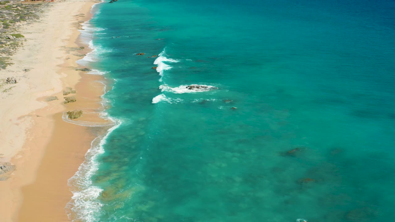 El Cardoncito shoreline from an elevated angle, with a larger white wave breaking around dark offshore rocks, pale beach to the left, and no visible people.
