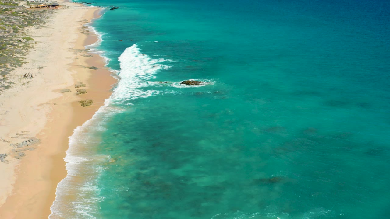 Aerial frame over El Cardoncito, Baja California Sur showing pale sand, turquoise shallows, a breaking wave line, scattered rocks offshore, low coastal vegetation, and no visible people.