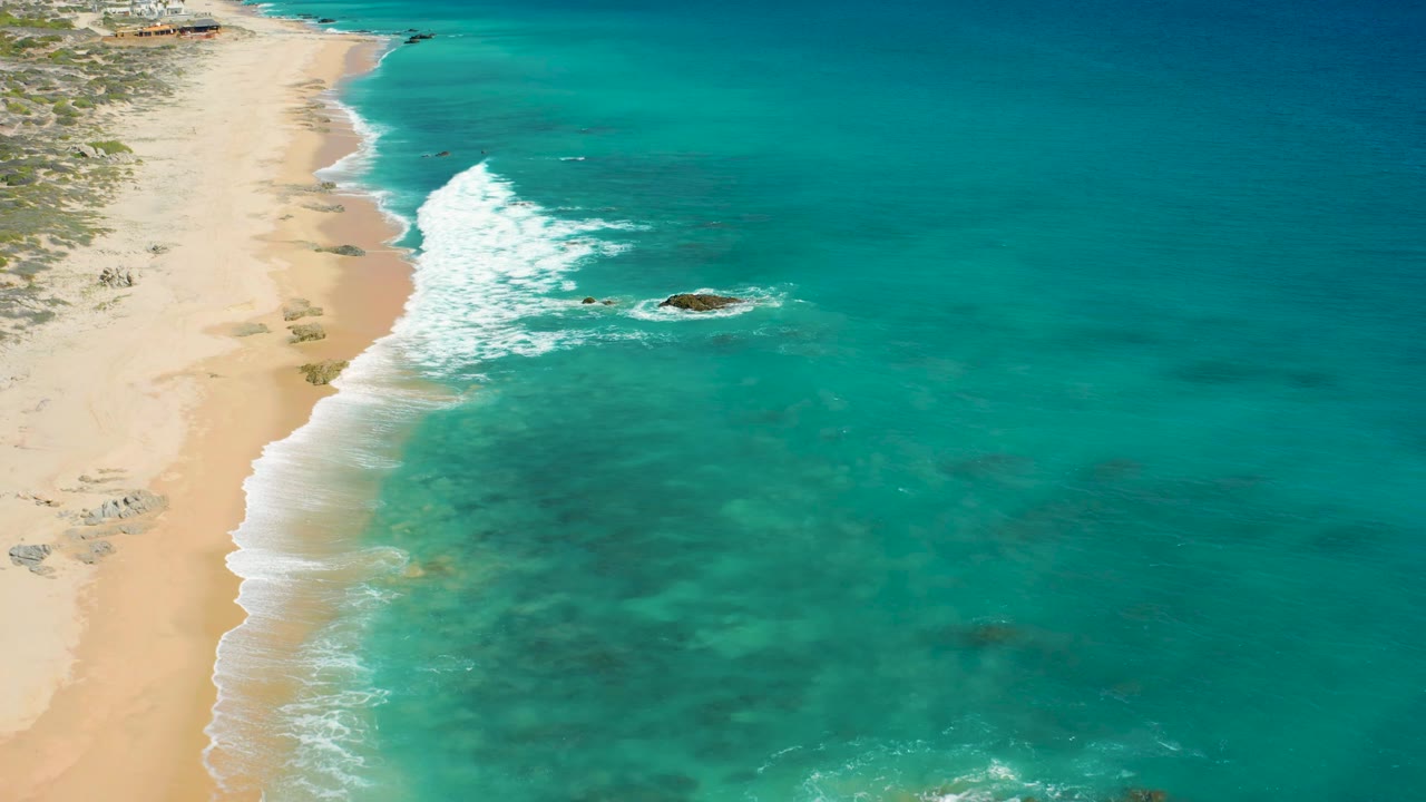 Aerial frame over El Cardoncito showing a diagonal strip of pale beach, white foam rolling ashore, a darker rock patch in the water, mottled shallow seabed tones, and no visible people.