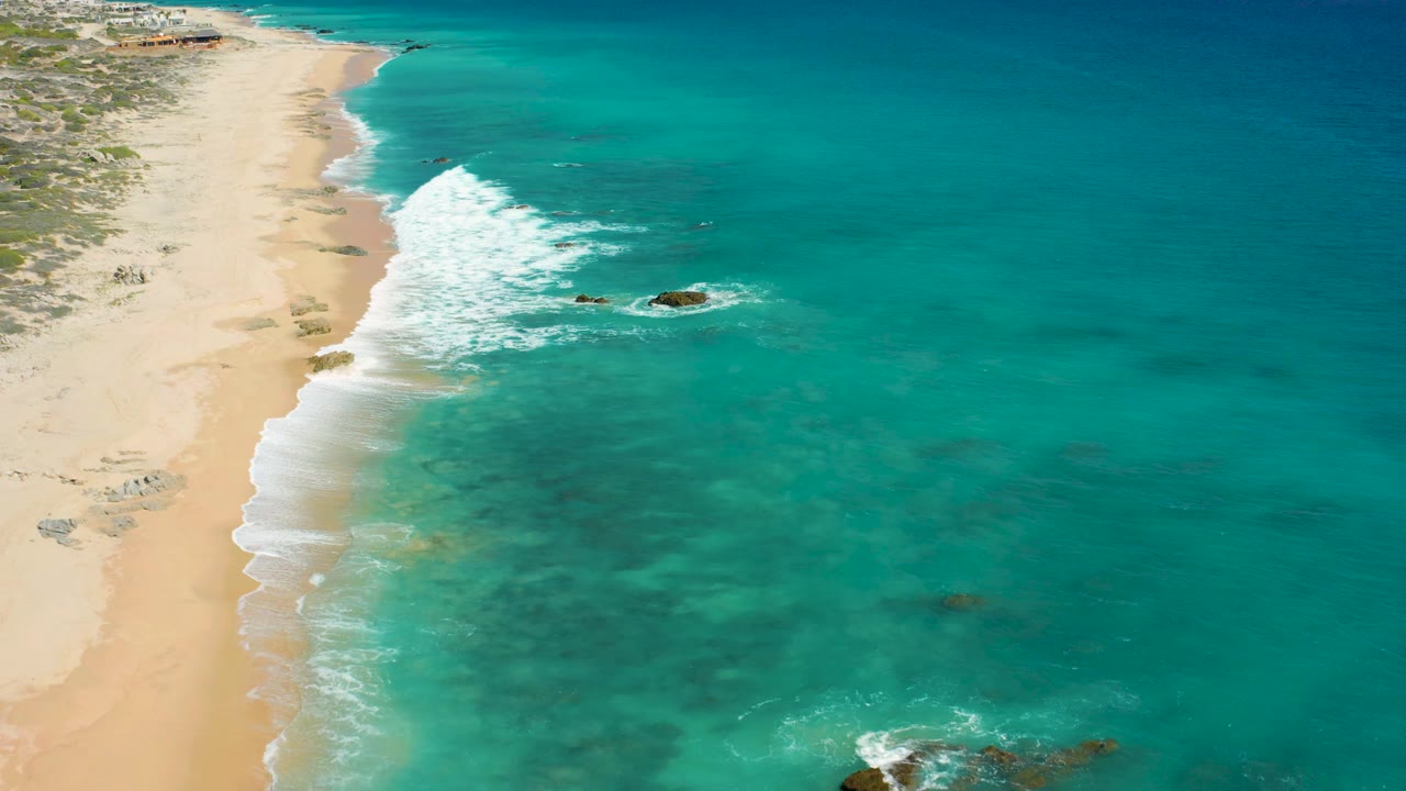 Aerial frame over El Cardoncito with the surf line advancing along the beach, a prominent offshore rock near center-right, shallow turquoise water, sandy coast, sparse vegetation, and no visible people.