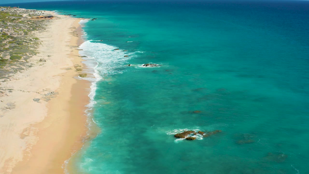 Opening aerial frame over El Cardoncito, Baja California Sur, with pale sand on the left, clear turquoise water on the right, offshore rocks, and no visible people.