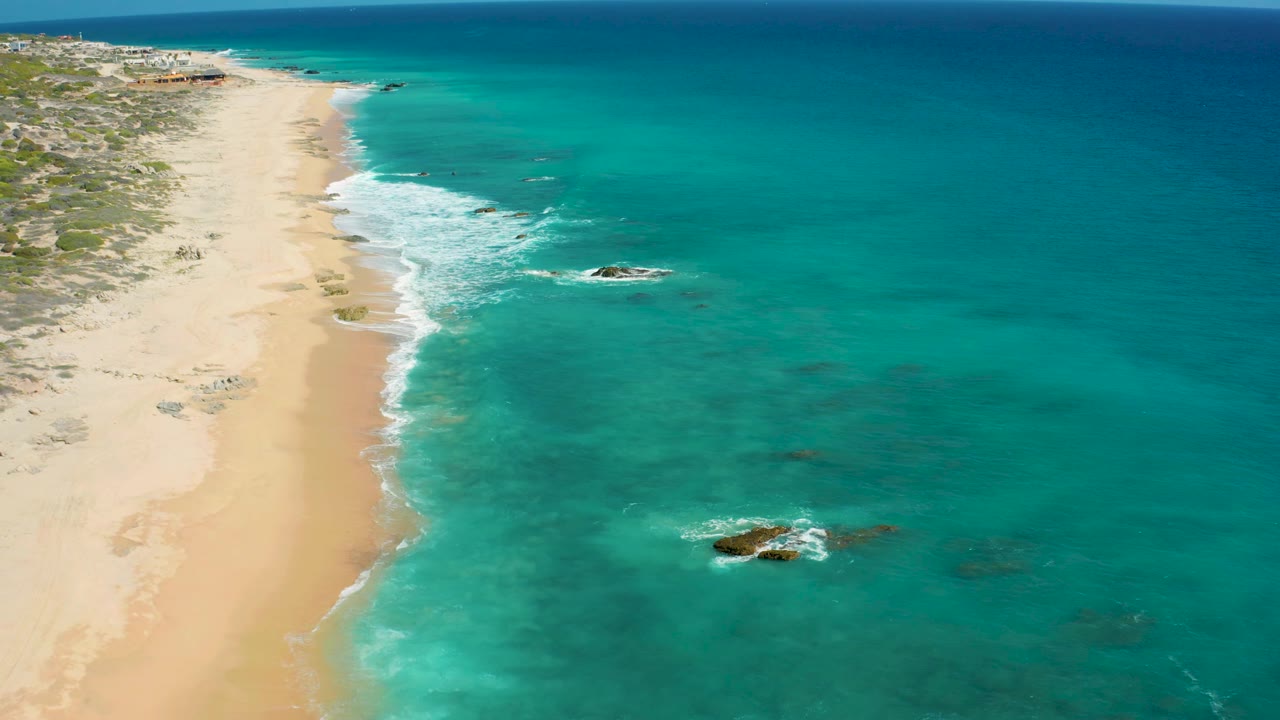 Elevated coastal frame in El Cardoncito with more open turquoise sea on the right, a narrow strip of pale beach on the left, scattered rocks, and no visible people.