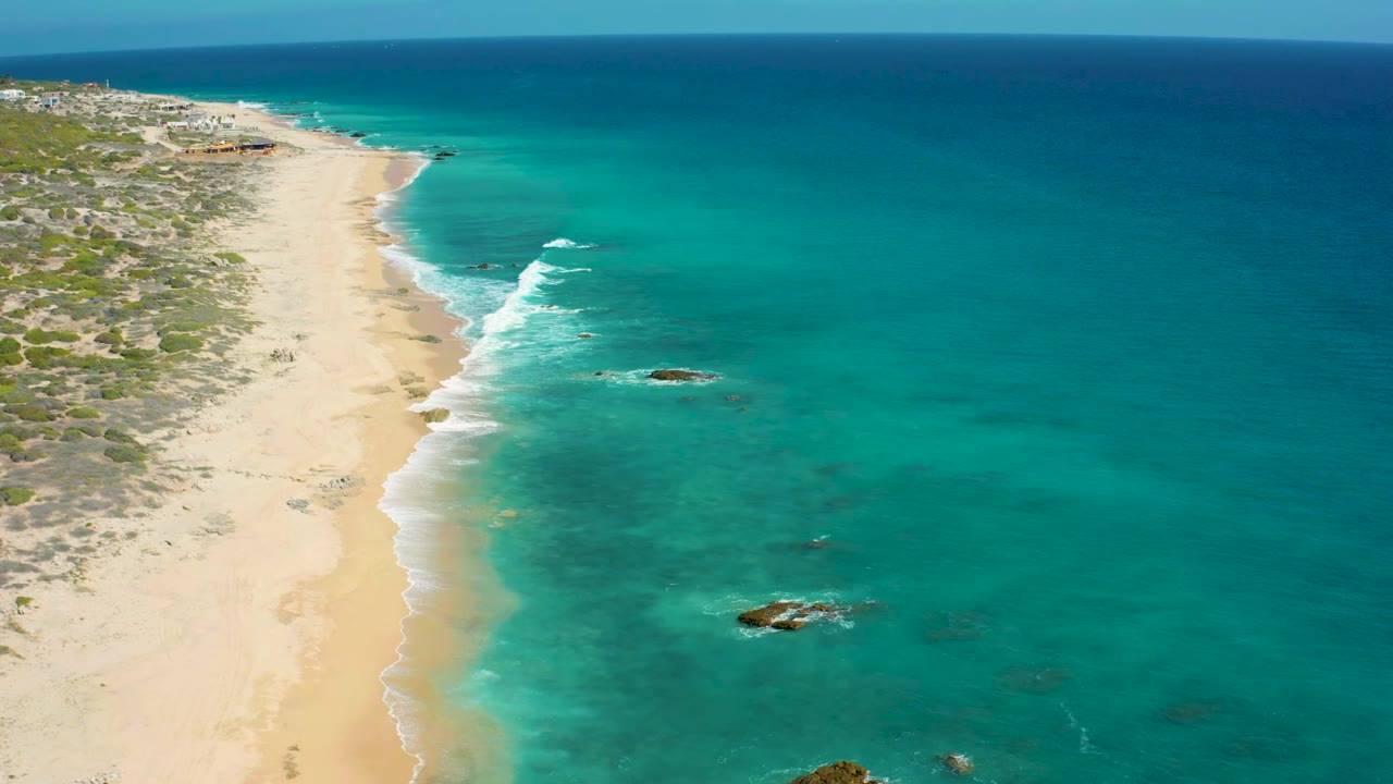 Aerial view of El Cardoncito with a bright surf line curving along the sand, rocky patches in clear water, and no visible people.