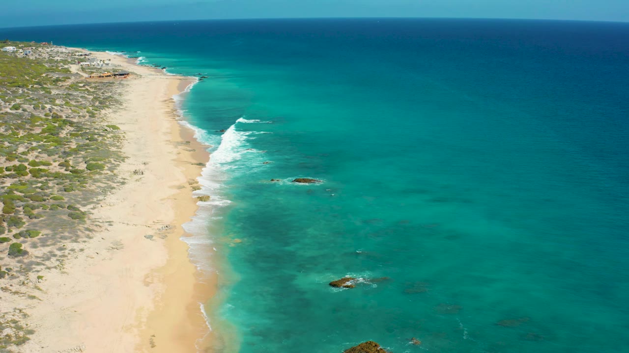 High view along El Cardoncito beach with long sand, foamy surf bands, clear water, low vegetation, and no visible people.