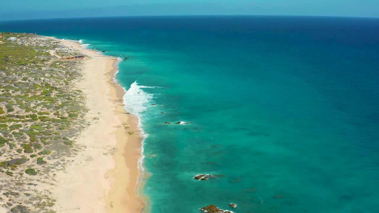 Aerial view over El Cardoncito with the sandy beach slightly left of center, white surf lines along the shore, dark blue water on the horizon, and no visible people.