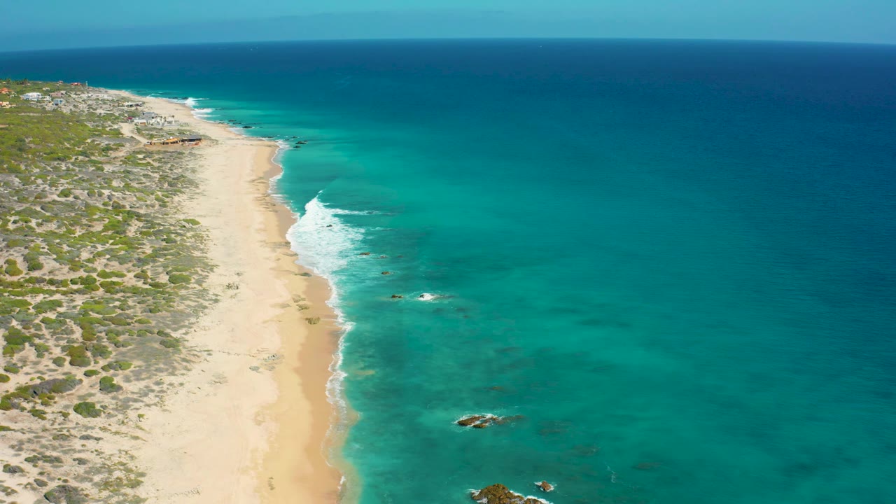 High aerial frame of El Cardoncito where scrubland borders the sand, turquoise water fills the right side, small waves break near shore, and no visible people appear.