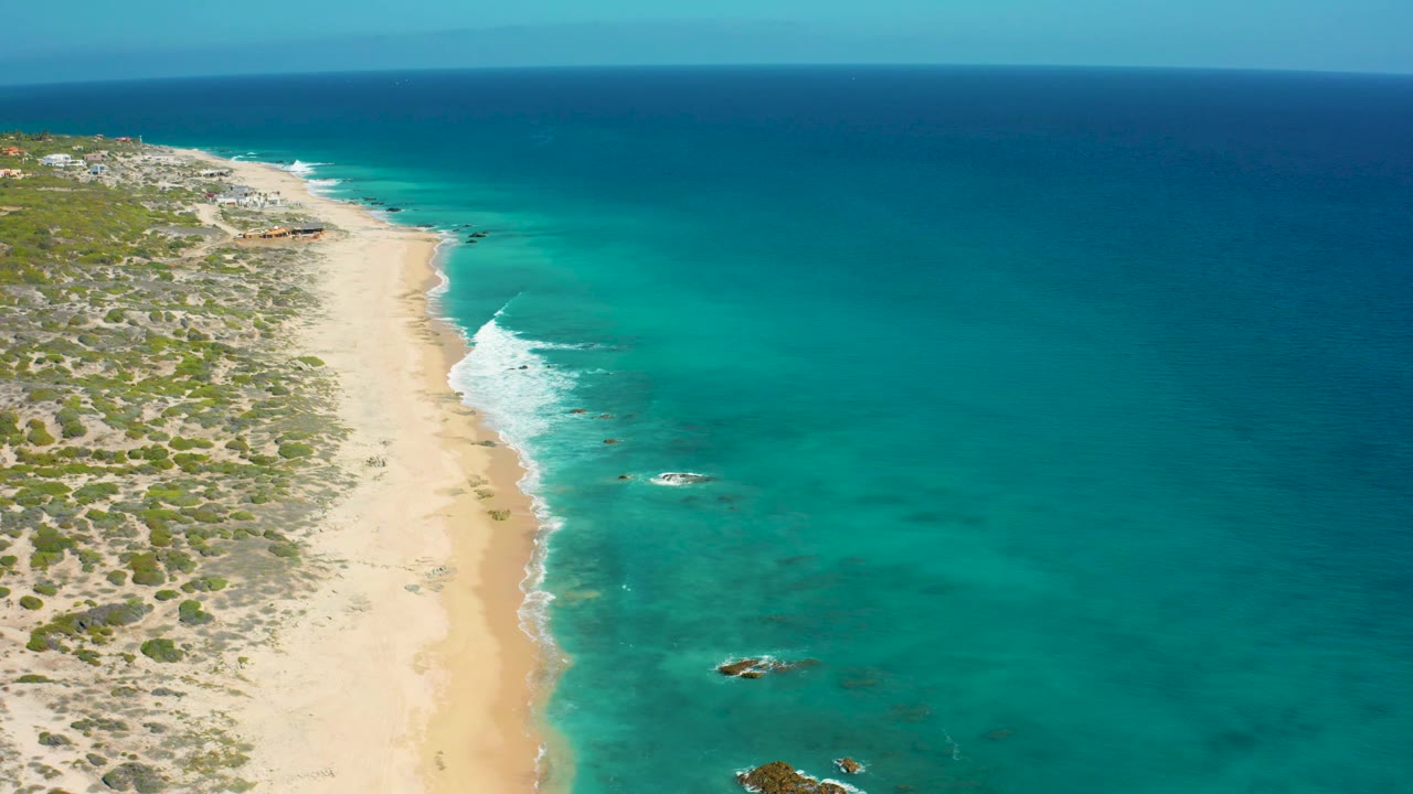 Aerial frame over El Cardoncito showing empty pale sand, low vegetation inland, a narrow white surf edge, open turquoise water, and no visible people.