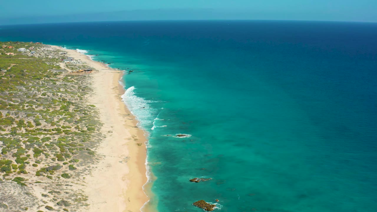 Elevated aerial frame over El Cardoncito shoreline, with the beach running diagonally between low scrub and blue-green water, plus no visible people on the sand or in the sea.