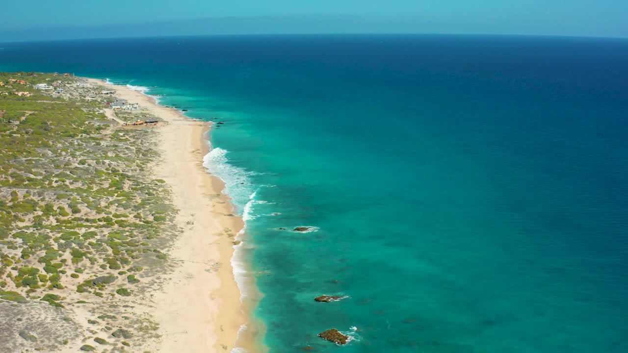 Aerial frame from the Baja California Sur coast showing pale beach below low green scrub, a band of turquoise shallows, deeper blue water beyond, and no visible people.