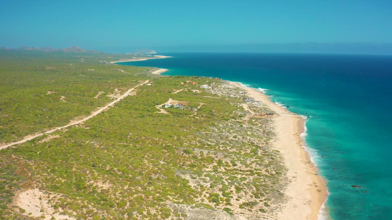 Aerial view over El Cardoncito, Baja California Sur, with green coastal scrubland, a pale beach, turquoise nearshore water, deep blue ocean beyond, and no visible people.