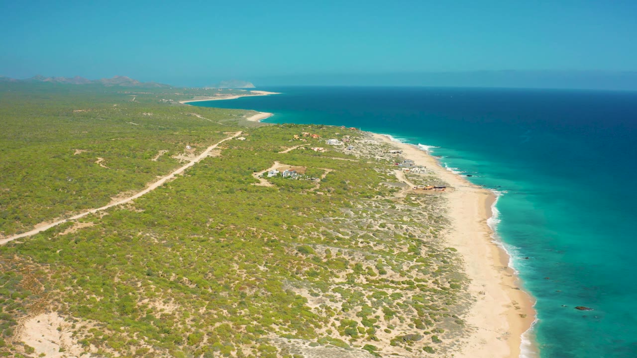 Wide aerial frame over El Cardoncito with calm blue water on the right, pale sand below, scrub-covered land on the left, distant coast on the horizon, and no visible people.