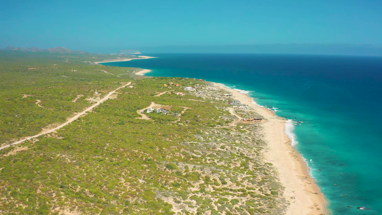 High coastal view with pale sand, blue sea, scrubland, dirt roads, and small structures near shore.